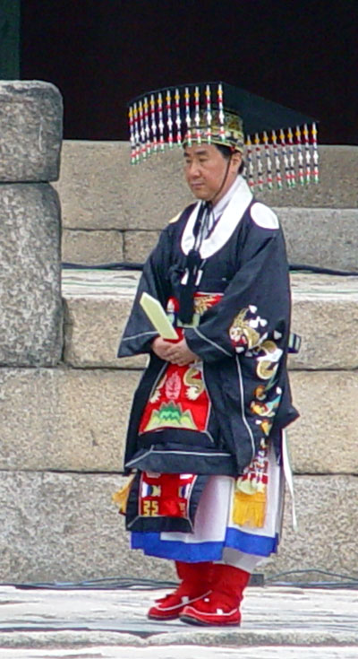 A male-presenting person in late middle age wearing a dark square hat with a row of colorful ornaments hanging from the front and back brim and an dark blue-grey robe with embroidery and a red panel in front, with a white robe underneath and red boots. He holds a long white piece of wood at waist level, pointing it upward.
